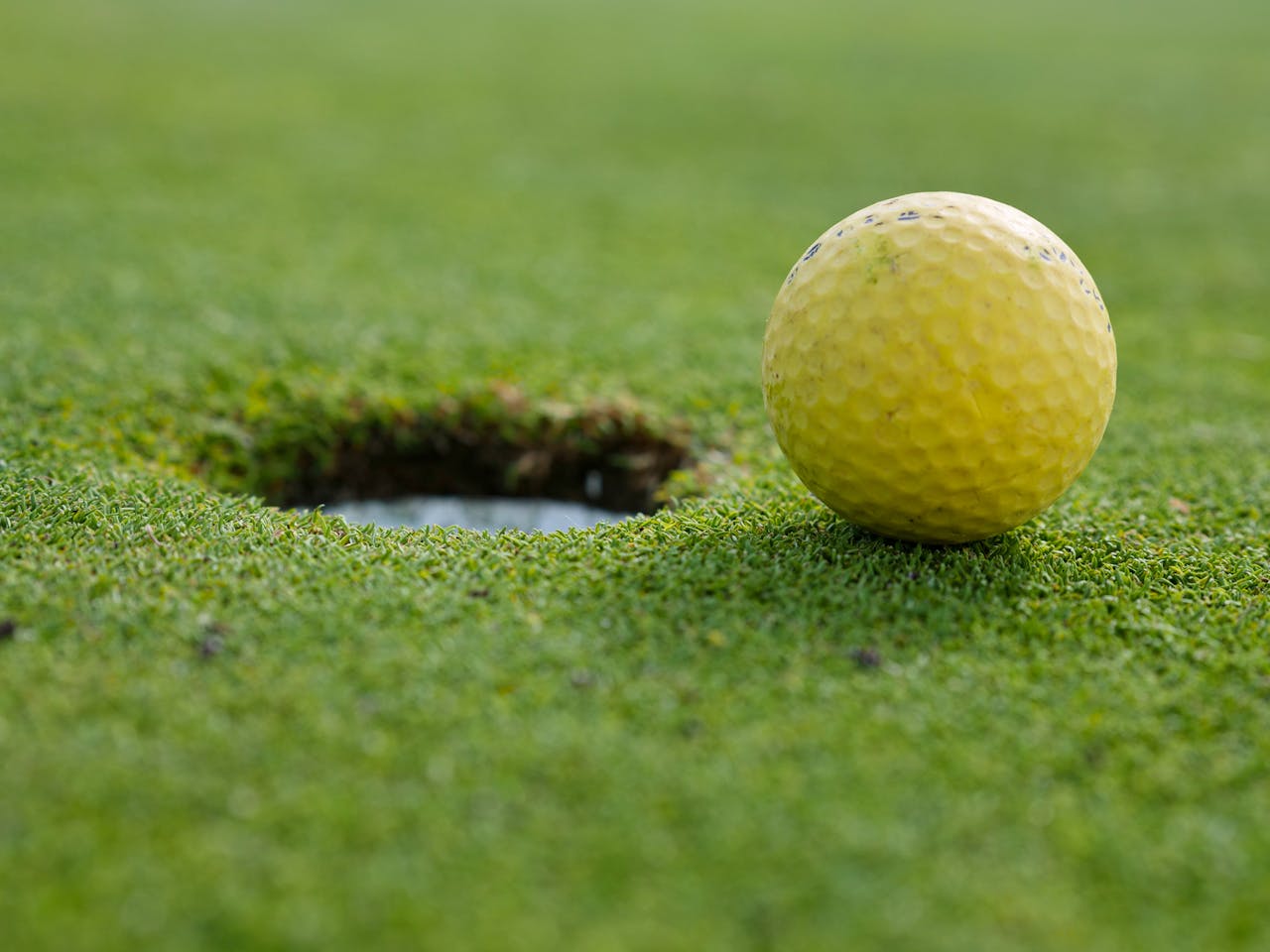 contact-img A yellow golf ball close to the hole on a grassy golf course.