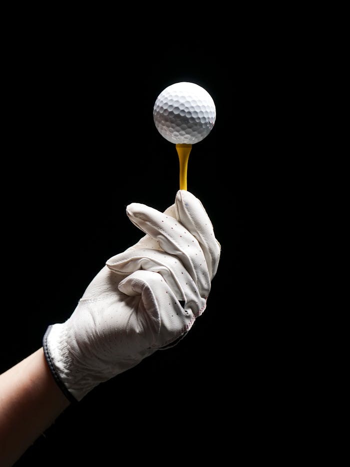 about-02 A gloved hand elegantly holds a golf ball on a tee against a black background.